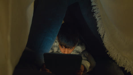 Nahaufnahme-Eines-Kleinen-Jungen-In-Einem-Selbstgebauten-Lager-Aus-Kissen,-Der-Nachts-Mit-Einem-Tablet-PC-Spielt